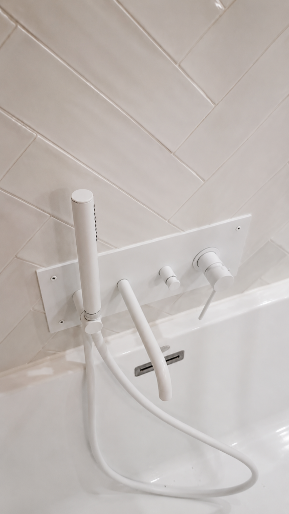 White wall-mounted bathtub faucet with a handheld shower, two control knobs, and a hose against white tiled wall (bath/shower fixture).