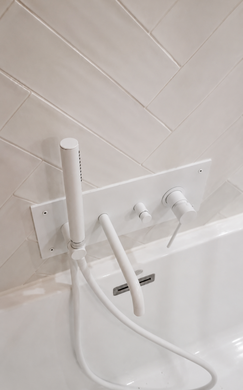 White wall-mounted bathtub faucet with a handheld shower, two control knobs, and a hose against white tiled wall (bath/shower fixture).